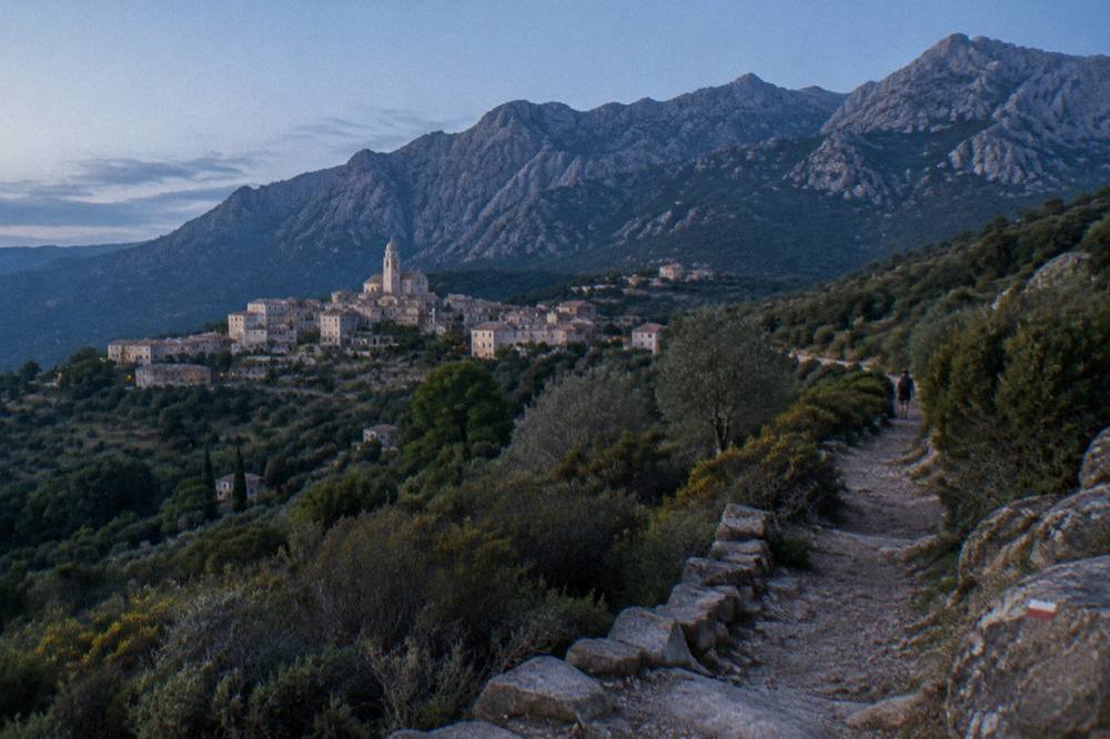 A marked trail winding through maquis shrubland toward a distant stone village