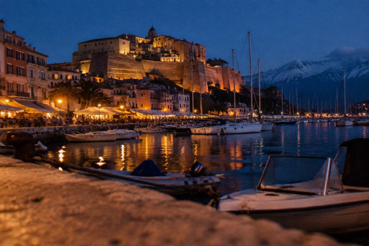 A Corsican harbour at dusk with boats moored along the quay and warm lights along the waterfront