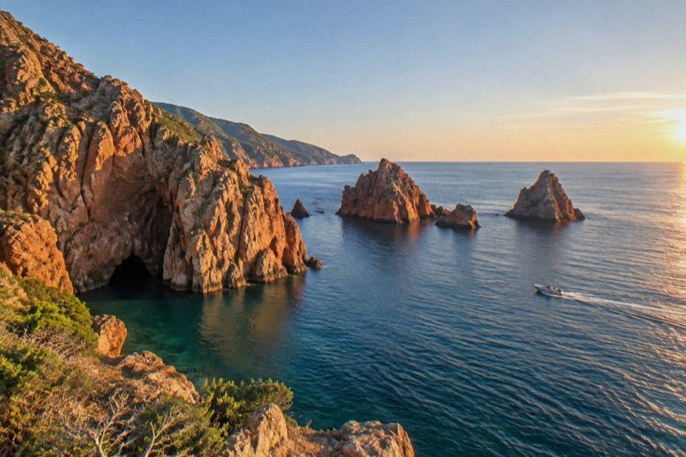 A small boat cruising past red rock islets at sunset off a Mediterranean coast