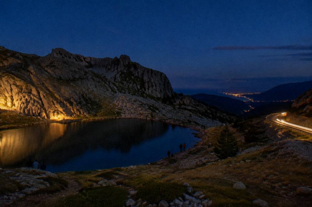 A car headlight trail on a winding mountain road at night with distant coastal lights