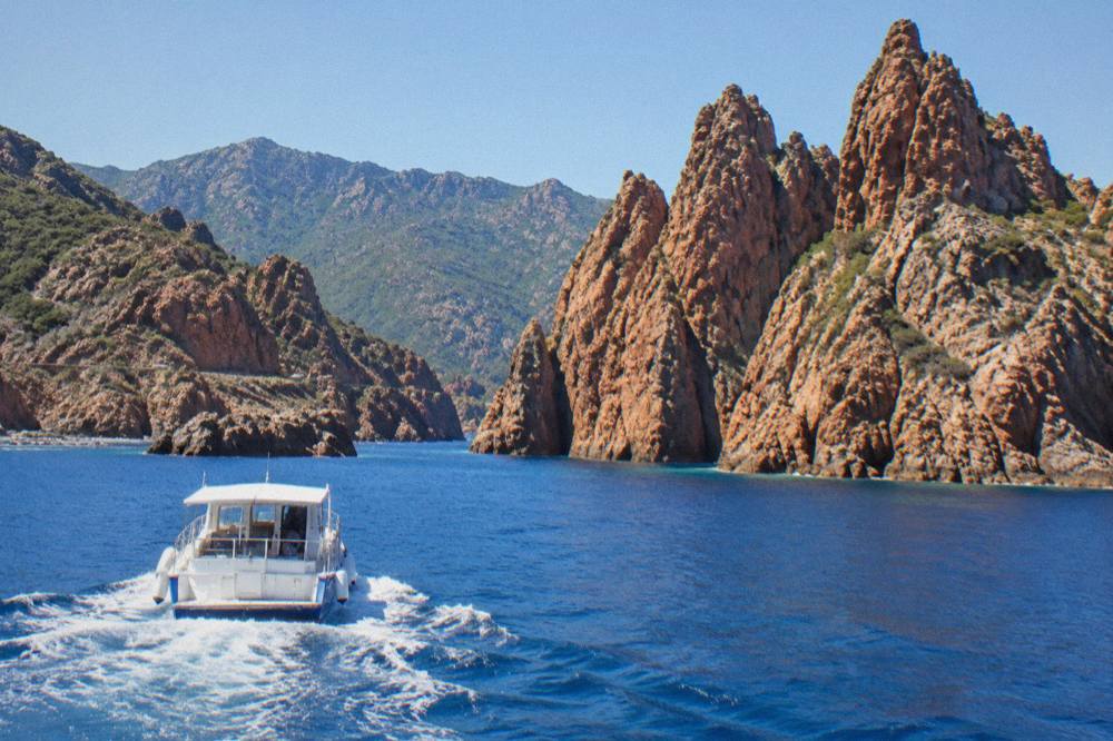 A boat crossing clear water toward the red rock formations of the Scandola Reserve