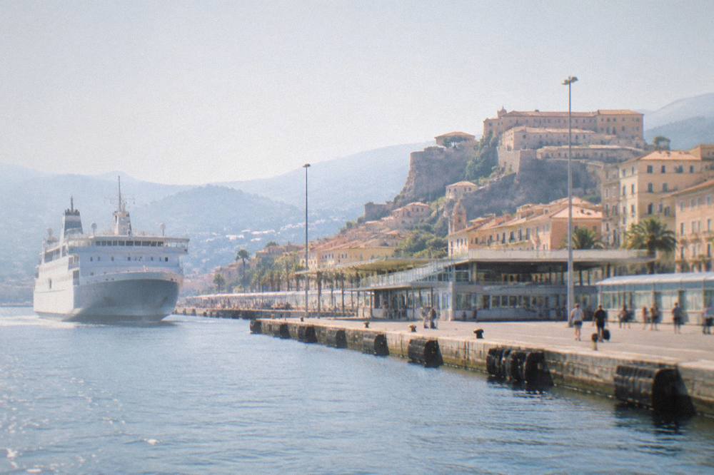 A ferry approaching a Corsican port town with a citadel above the water