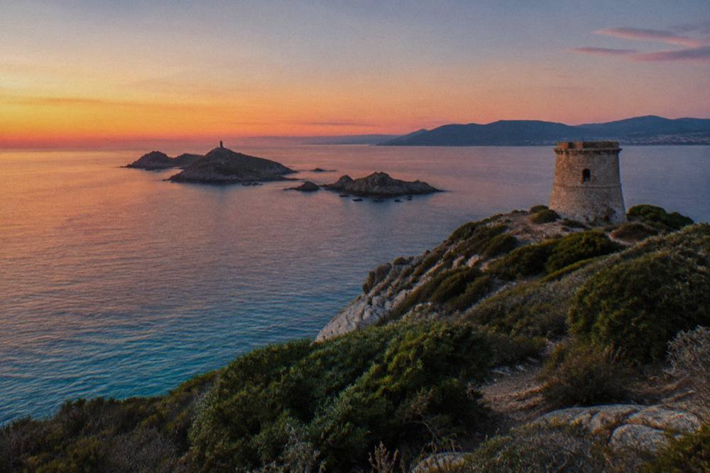 A stone watchtower on a headland above turquoise water at sunset in Corsica