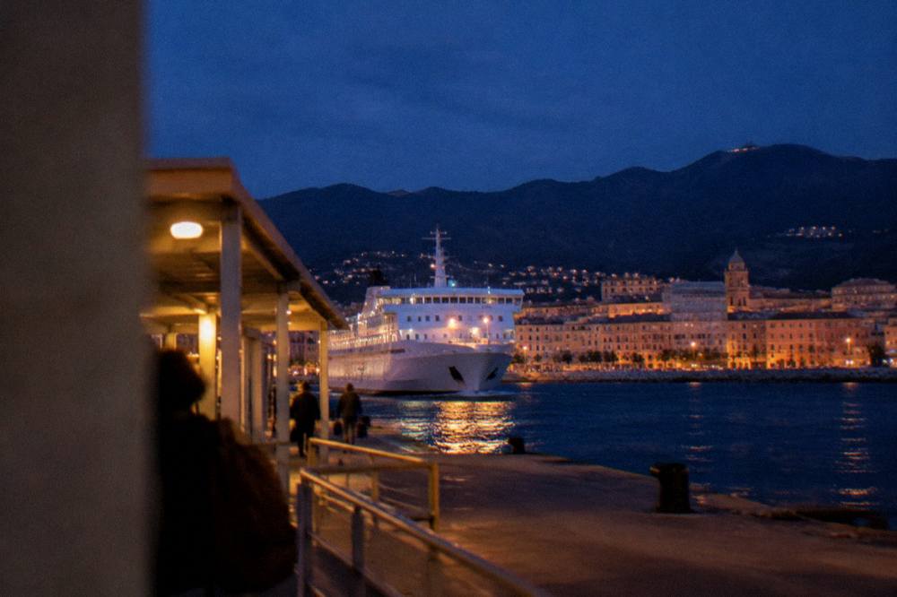 A white ferry approaching a Corsican harbour town at dusk with mountains behind