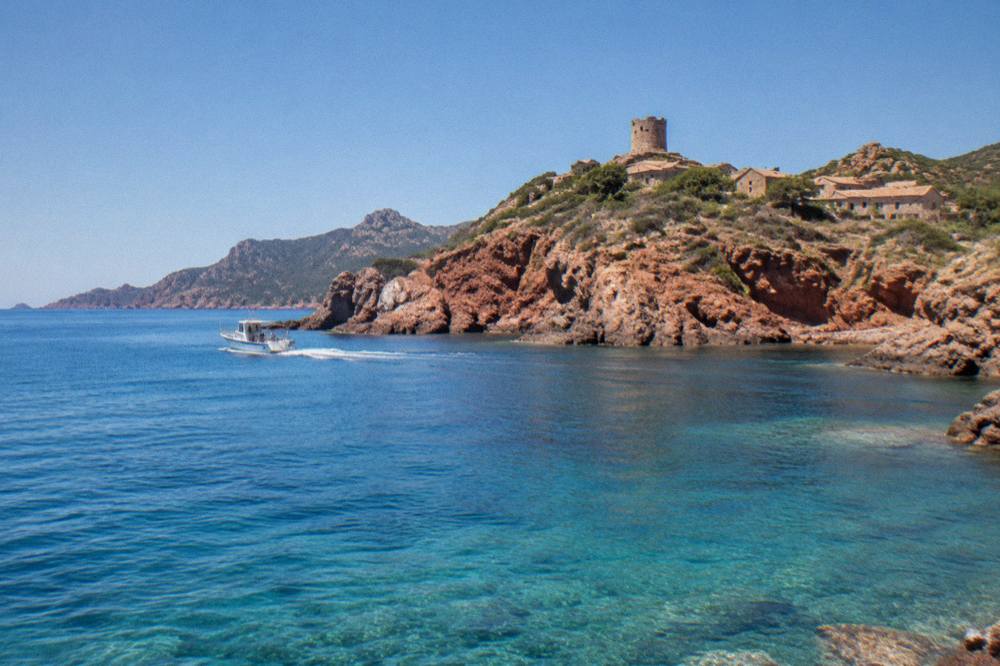 A small boat cruising along red cliffs above clear turquoise water on a bright calm day