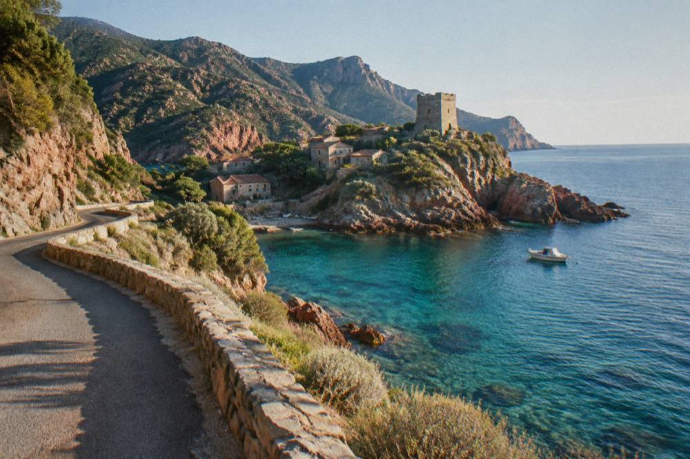 A coastal road winding above a turquoise cove with a small boat anchored offshore