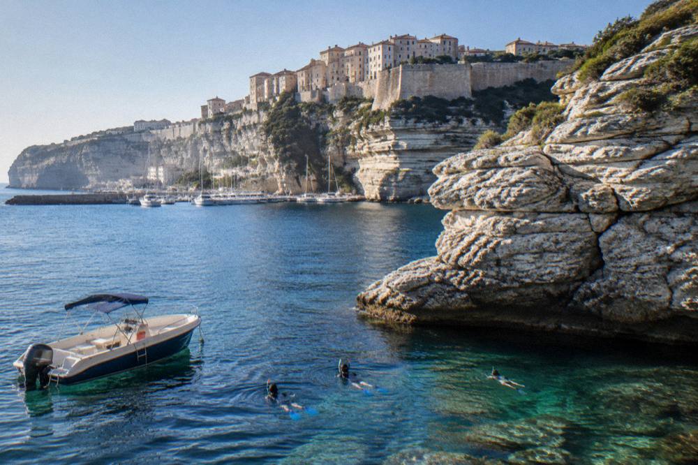 Snorkellers in the water beside a small boat near a rocky Corsican headland
