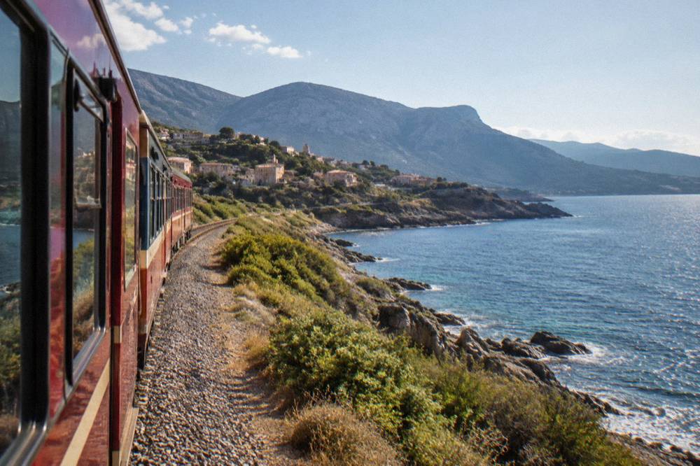 A red Corsican train on a coastal rail line beside the sea in the Balagne