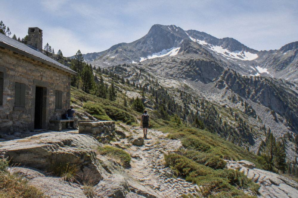 A stone refuge building on a mountain slope with a hiker resting outside