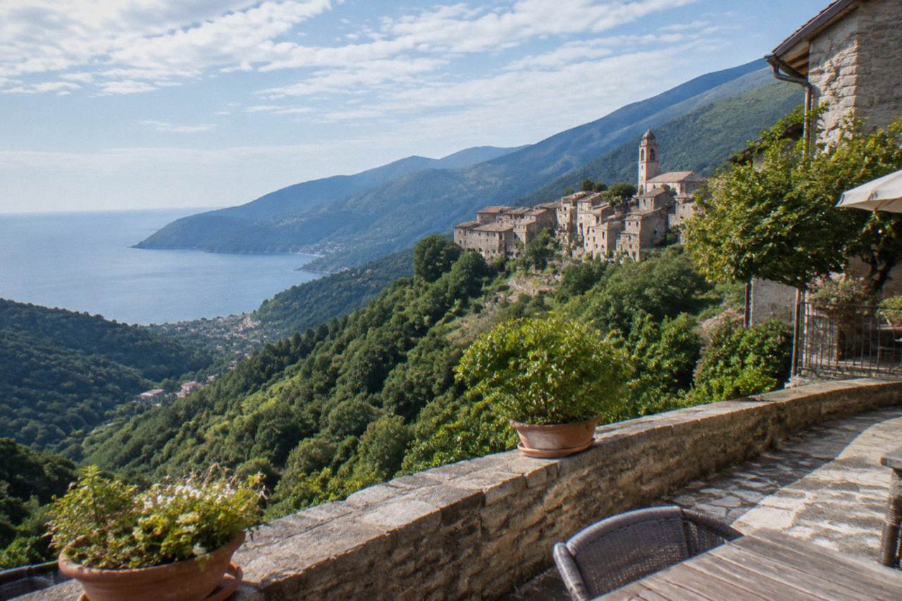 A stone hotel terrace above a calm Mediterranean bay with wooded hills in the background