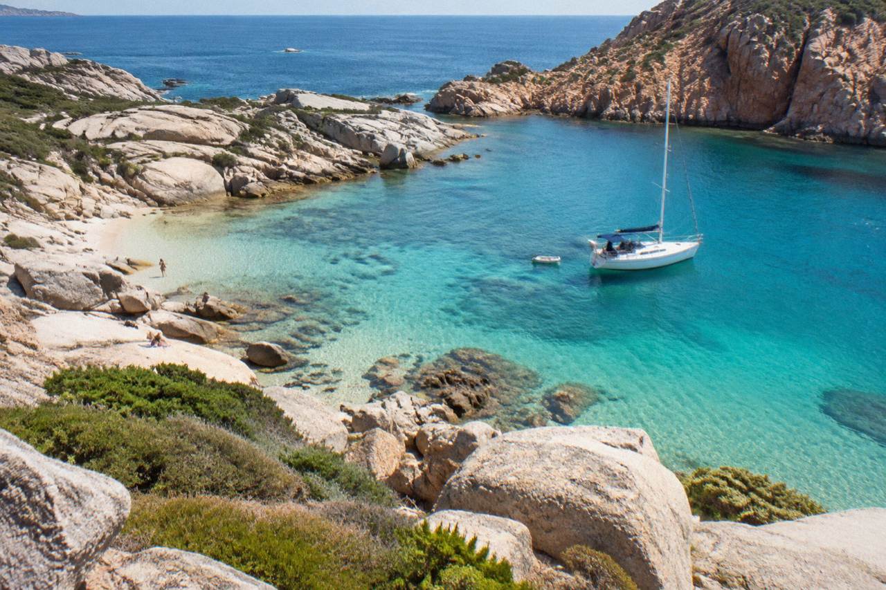 A sailing boat anchored in a calm cove below red granite cliffs on the Corsican coast