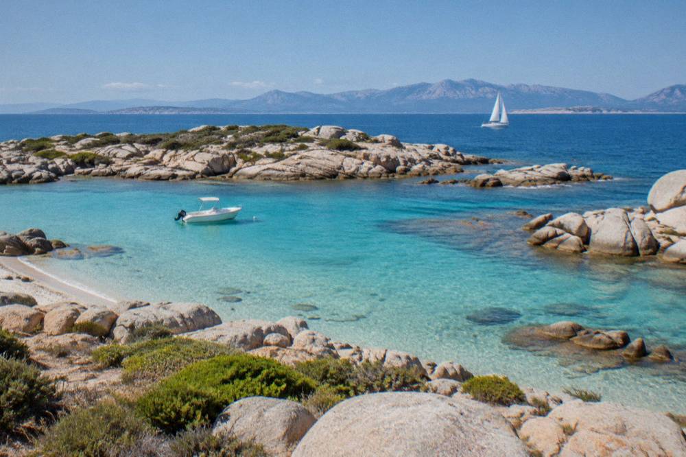 A yacht under sail in blue water with the Corsican mountains faint on the horizon