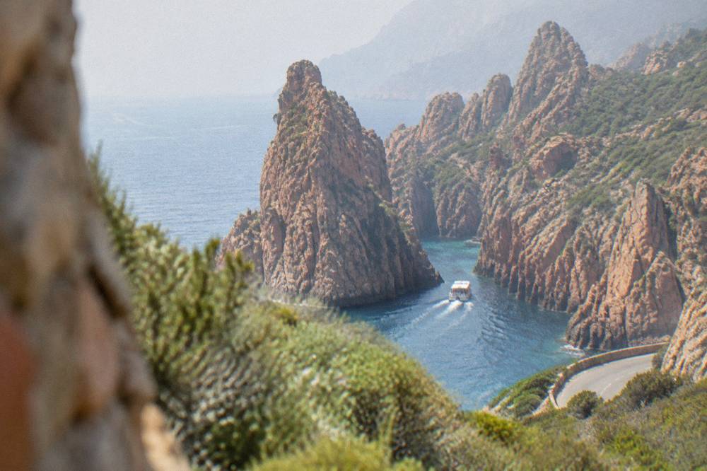 A small boat entering a red rock inlet between the Calanques de Piana