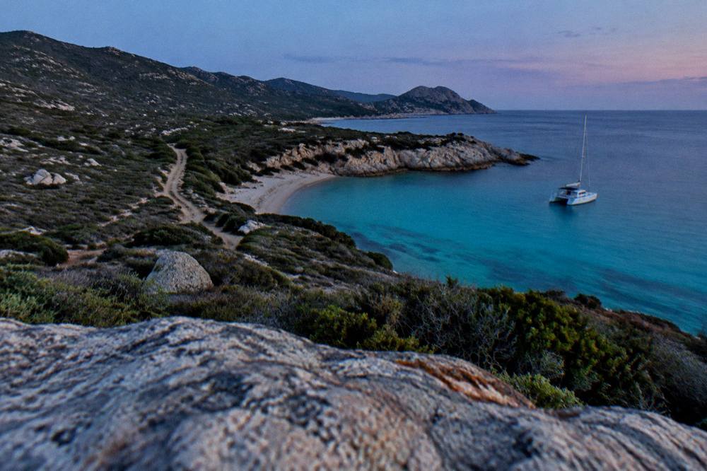 A catamaran moored in turquoise water near a sandy cove in the Agriates