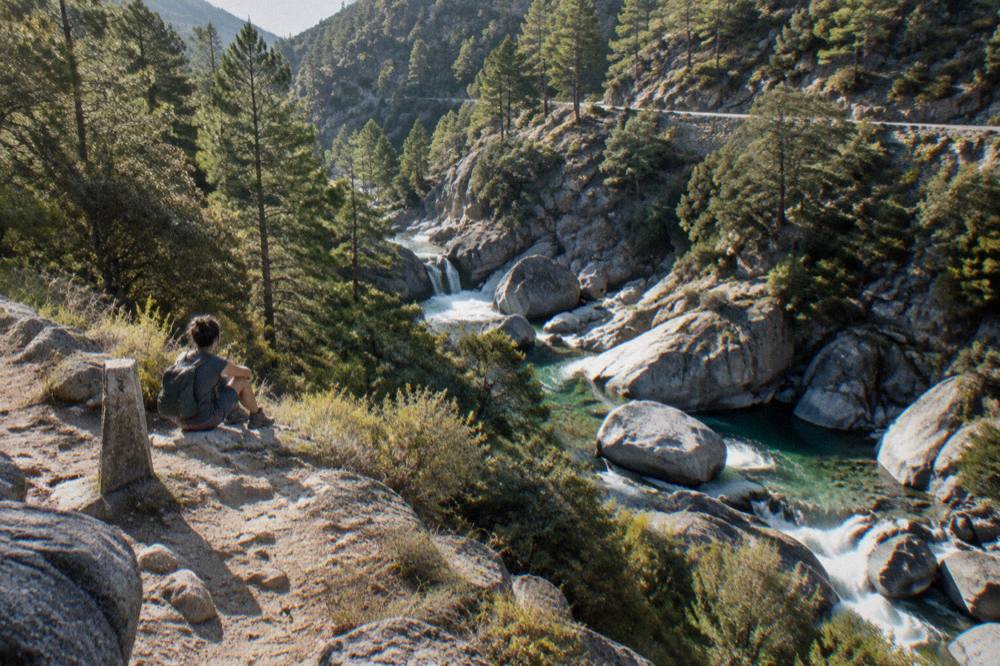 A lone hiker with a small backpack resting beside a marker post on a rocky Corsican mountain trail