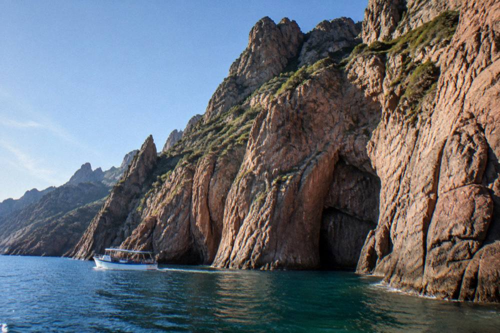 A small passenger boat cruising close to red granite cliffs on the Corsican west coast