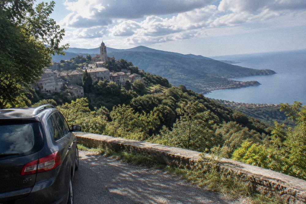 A family car parked beside a narrow mountain road with a view over a Corsican coastal bay