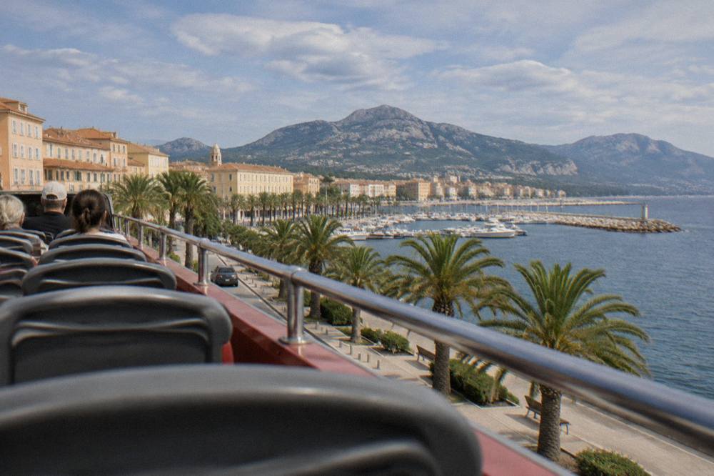 An open-top tour bus on a seaside road in Ajaccio with the gulf in the background