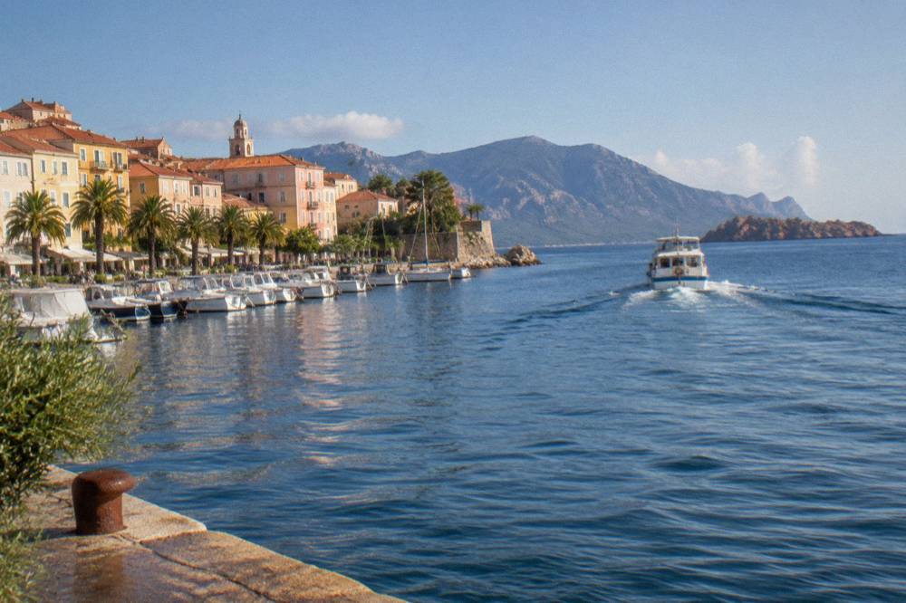 A small passenger boat heading out of Ajaccio harbour toward red granite islands on the horizon