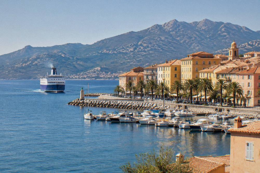 A ferry at sea approaching a Corsican port town in early morning light