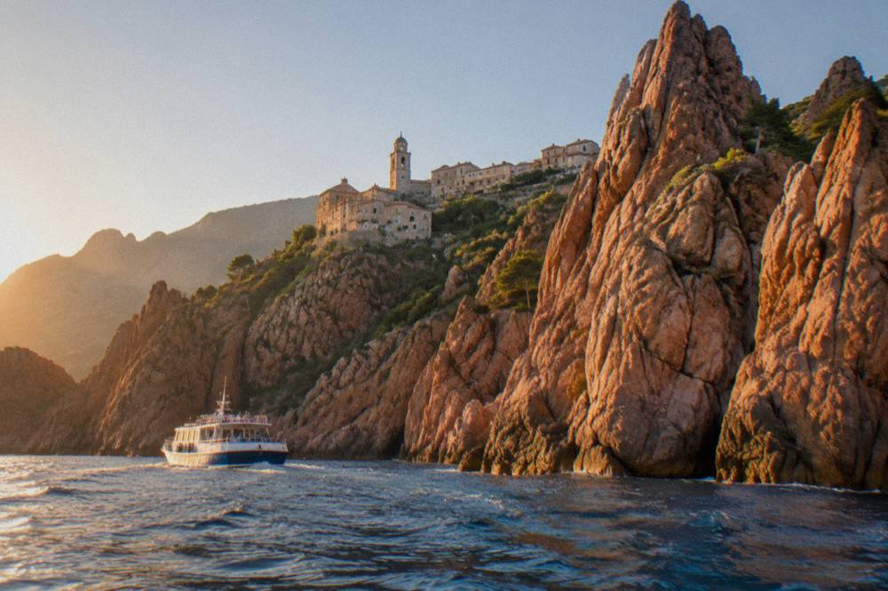 A small passenger boat passing close to weathered red rock towers on the Corsican coast near Piana