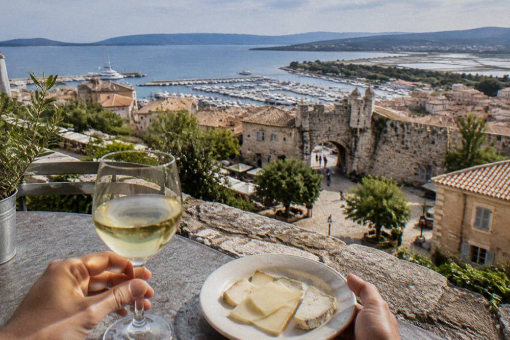 Hands holding a glass of white wine and a small plate of cheese at an outdoor tasting table