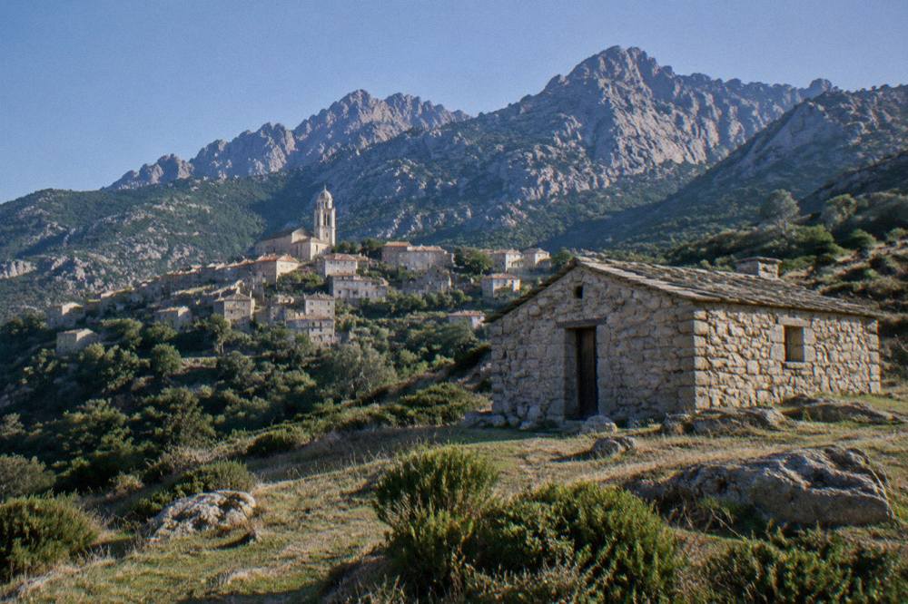 A stone refuge hut on a grassy plateau with mountain summits behind it