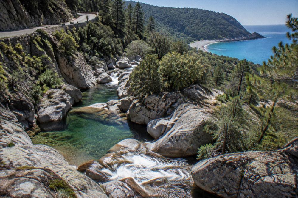 Turquoise sea and a pebble beach beneath wooded hills at the end of a coastal path in Corsica